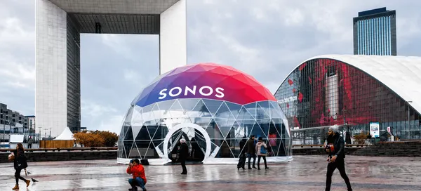 Transparent geodesic event dome with SONOS branding on its red and blue roof, installed in a modern urban plaza.
