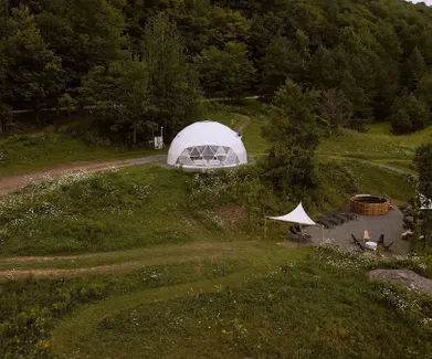 White geodesic glamping dome with an outdoor hot tub and seating area in a forested meadow.
