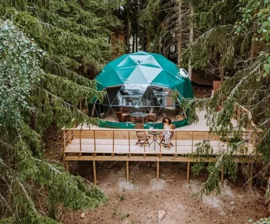 woman sitting in front of green geodesic dome on wooden deck in the forest