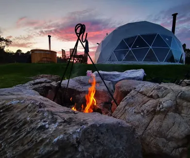 Geodesic glamping dome at sunset with an outdoor fire pit, wooden hot tub and dining area surrounded by nature.