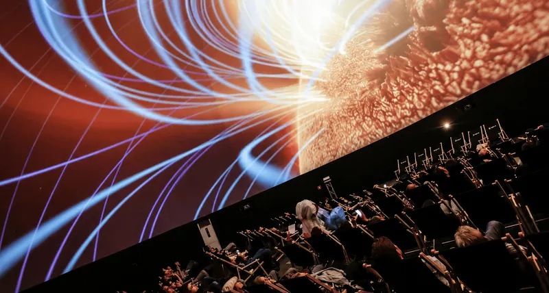 Seated audience watching an immersive dome projection featuring luminous blue light trails and orange abstract visuals.  Copy