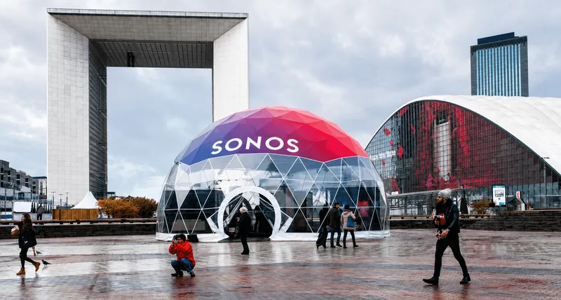 Transparent geodesic event dome with SONOS branding on its red and blue roof, installed in a modern urban plaza.