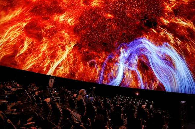 Audience seated inside a large immersive dome theatre beneath a curved projection screen displaying vivid red and blue abstract visuals.