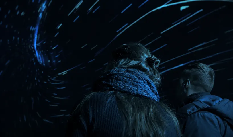Visitor watching a starfield projection inside a dark immersive dome, with streaks of blue light surrounding the viewing space.