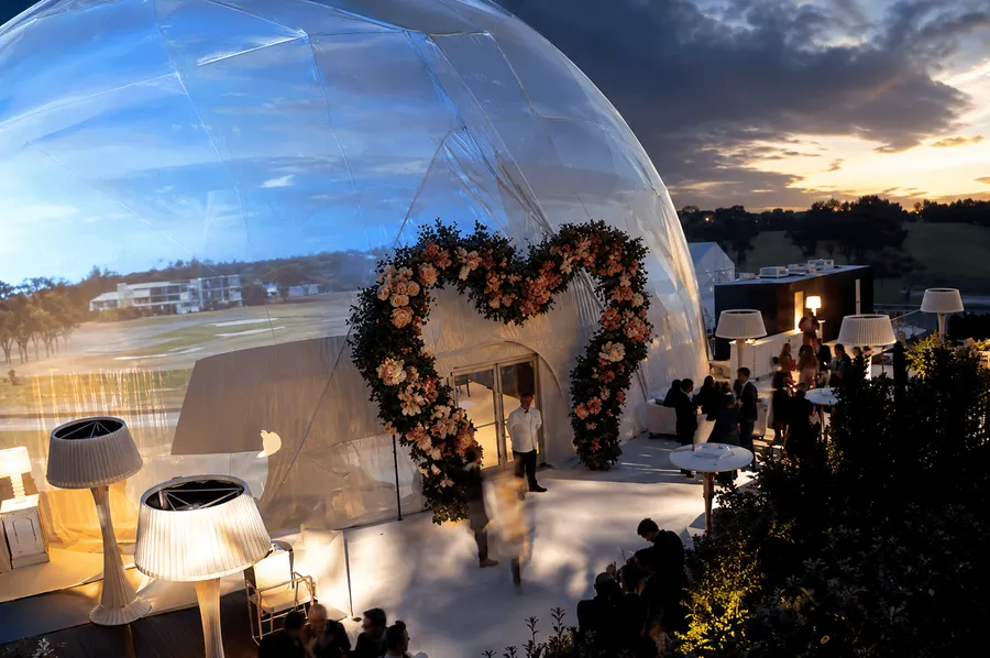 Geodesic event dome with a floral entrance arch, illuminated outdoor seating and guests attending an evening celebration.