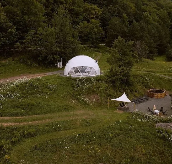 White geodesic glamping dome with an outdoor hot tub and seating area in a forested meadow.