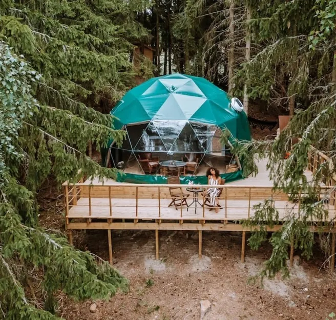woman sitting in front of green geodesic dome on wooden deck in the forest