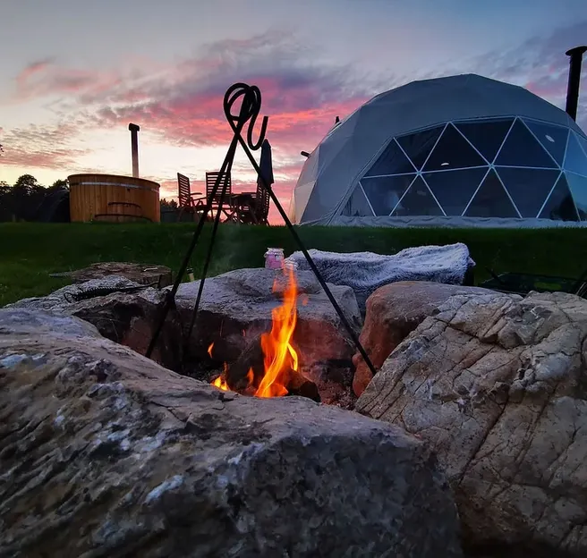 Geodesic glamping dome at sunset with an outdoor fire pit, wooden hot tub and dining area surrounded by nature.
