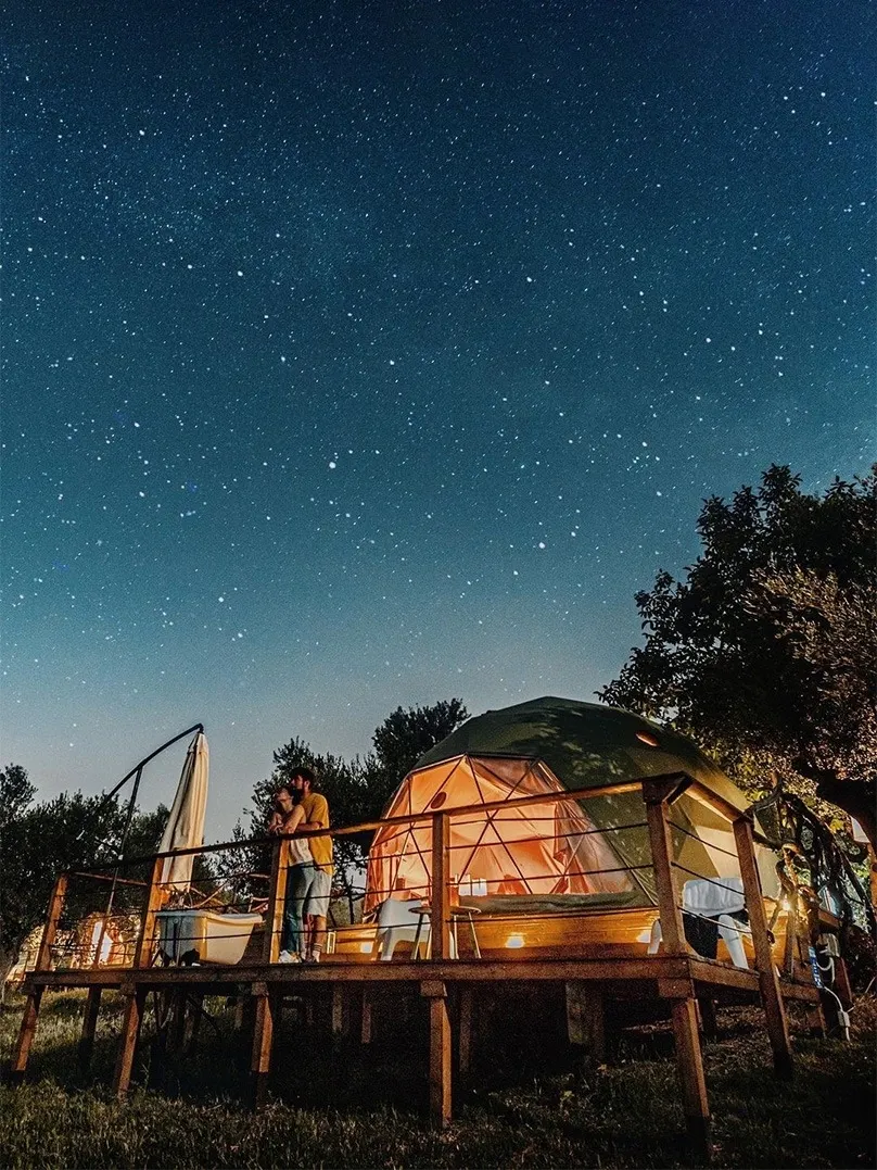 green geodesic dome on a wooden platform, in front of the dome a hugging couple admiring the starry sky