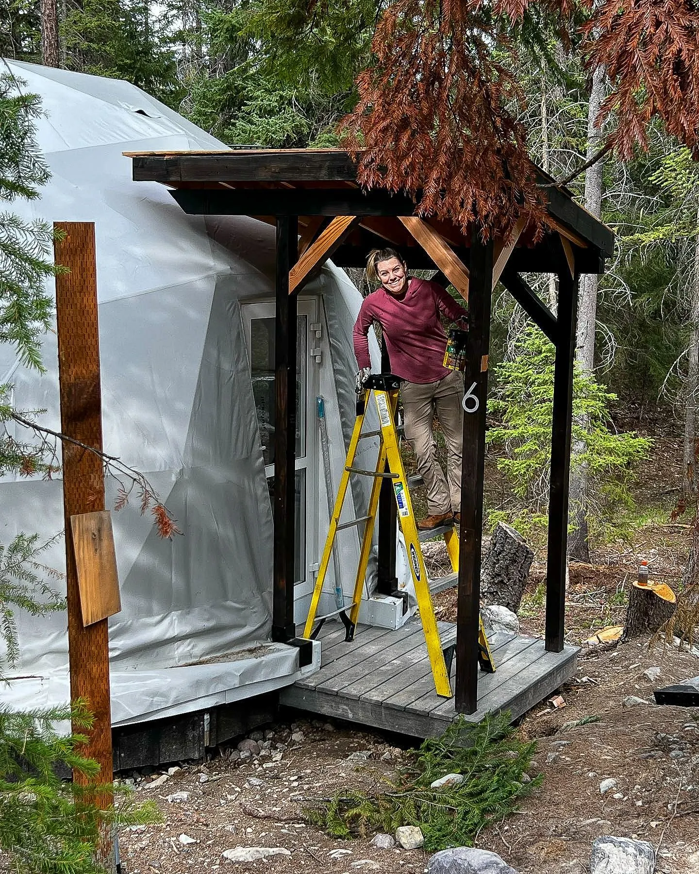 a woman standing on the ladder in front of white geodesic dome in the forest