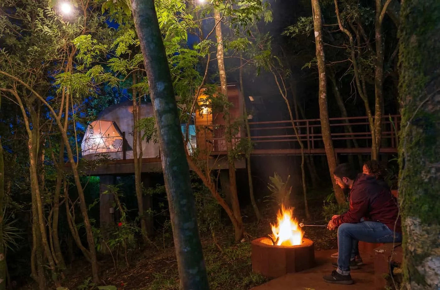 Guests sitting by a campfire beside an illuminated geodesic glamping dome in the forest at night.