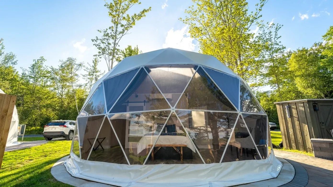 White geodesic glamping dome with a panoramic window, surrounded by trees and greenery.