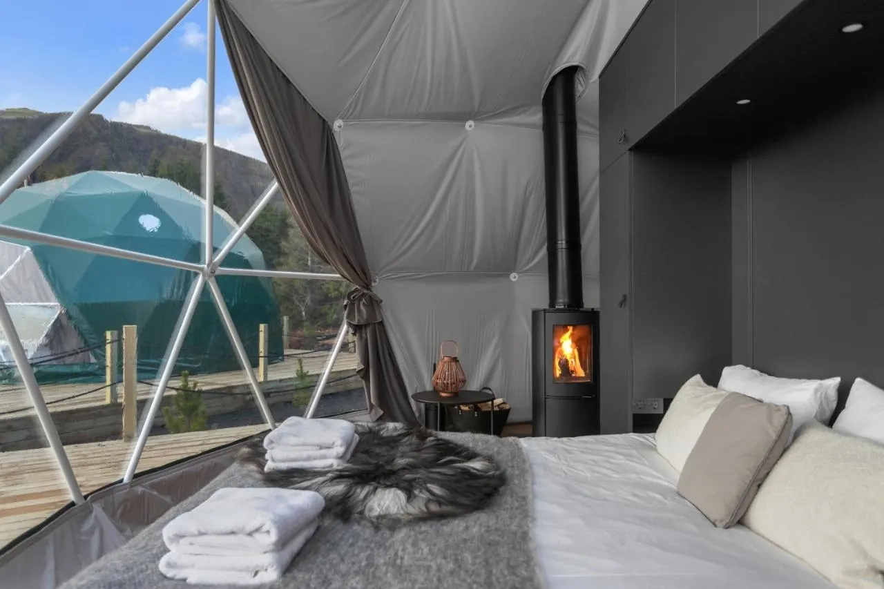 Interior of a geodesic glamping dome with a double bed, wood-burning stove and mountain view.