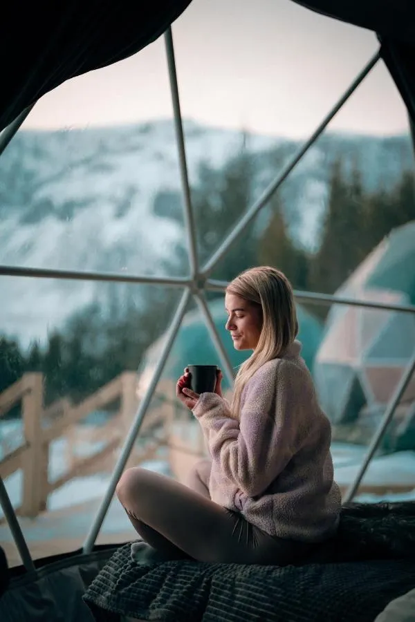 the interior of a geodesic dome with a view of the window, a woman is sitting on a bed with a cup of tea