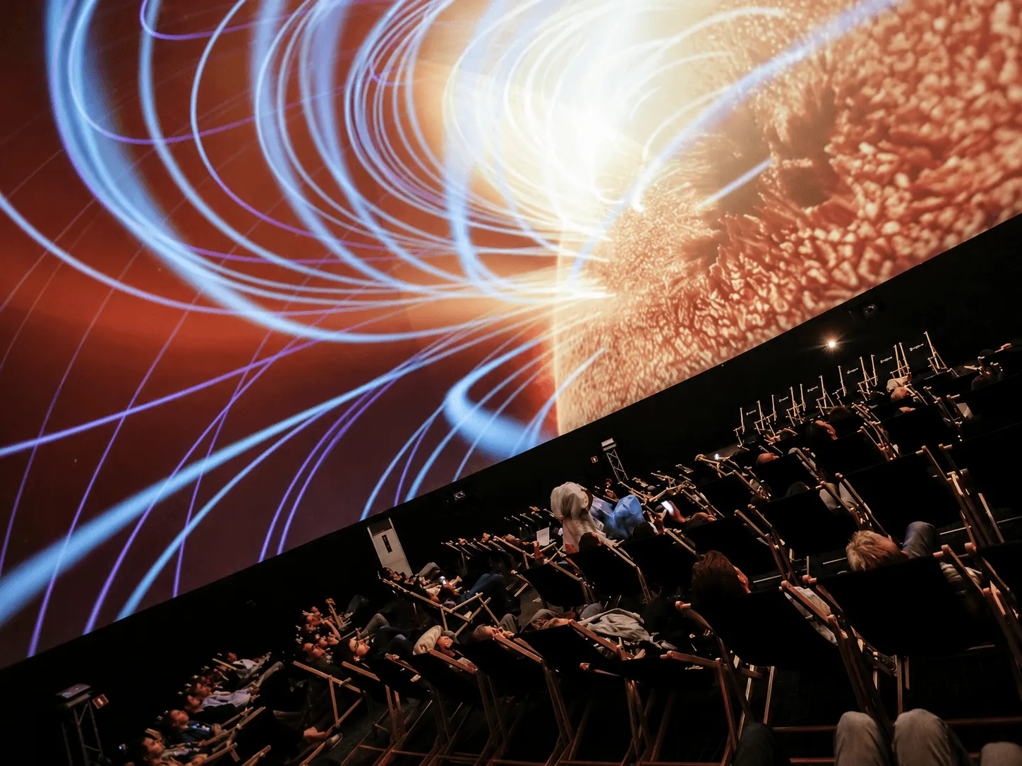 Seated audience watching an immersive dome projection featuring luminous blue light trails and orange abstract visuals.  Copy
